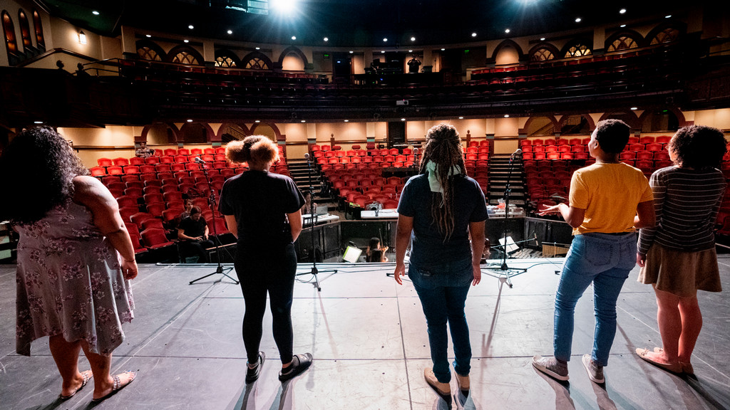 students stand on stage during rehearsal looking out to a large empty house