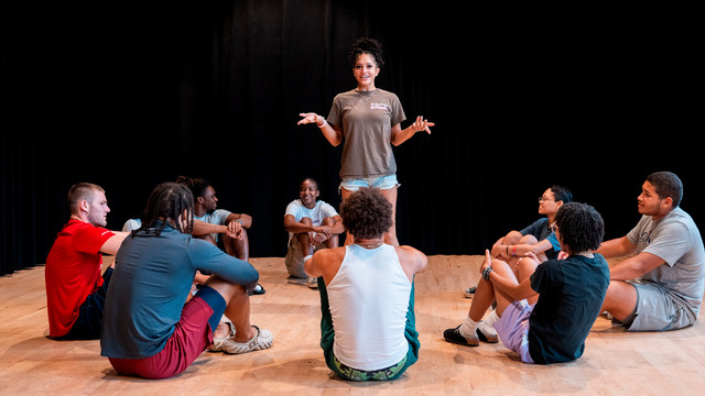 a group of theater students sit in a circle while one stands in the center