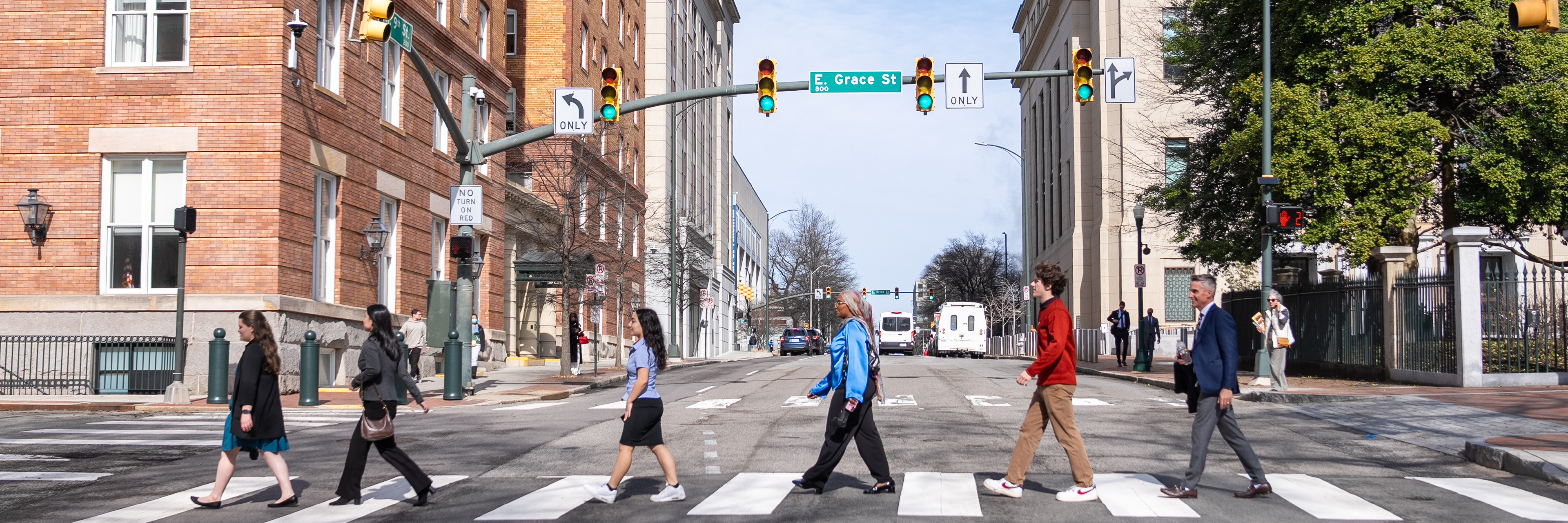 A group of students and alumni walking in a line through a crosswalk in downtown Richmond. A sign for Grace St. is above them and the image is reminiscent of the Beatles Abbey Road album cover.