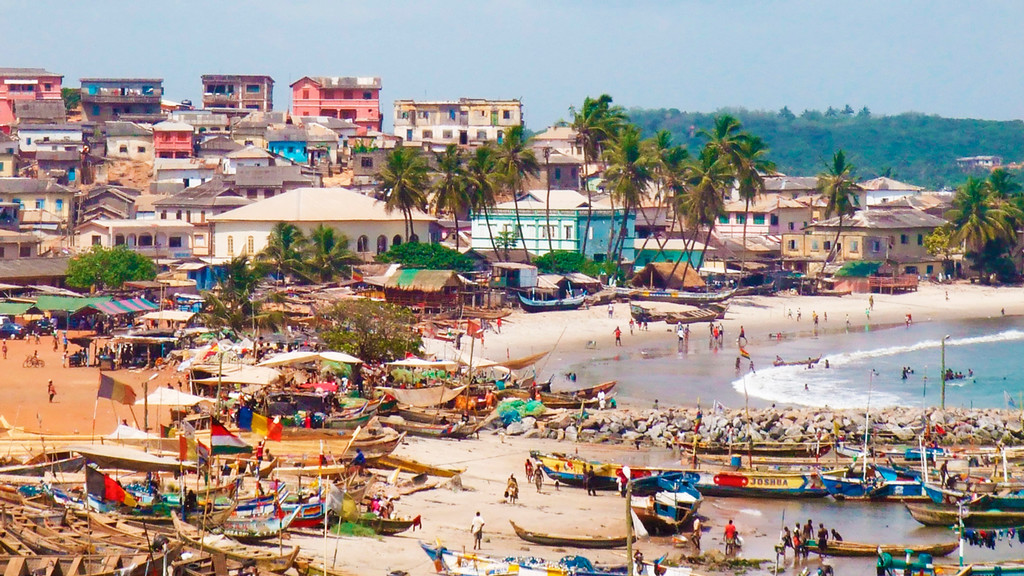 A busy beach full of people and fishing boats with mountains in the background