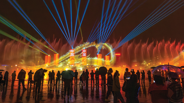 a crowd gathering at night to view a light show featuring a dragon against the city skyline