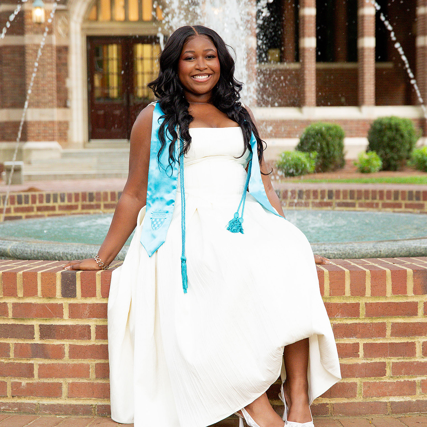 Layla Harmon sitting in front of a fountain on campus. She is wearing a white dress and her graduation stoles and smiling at the camera.