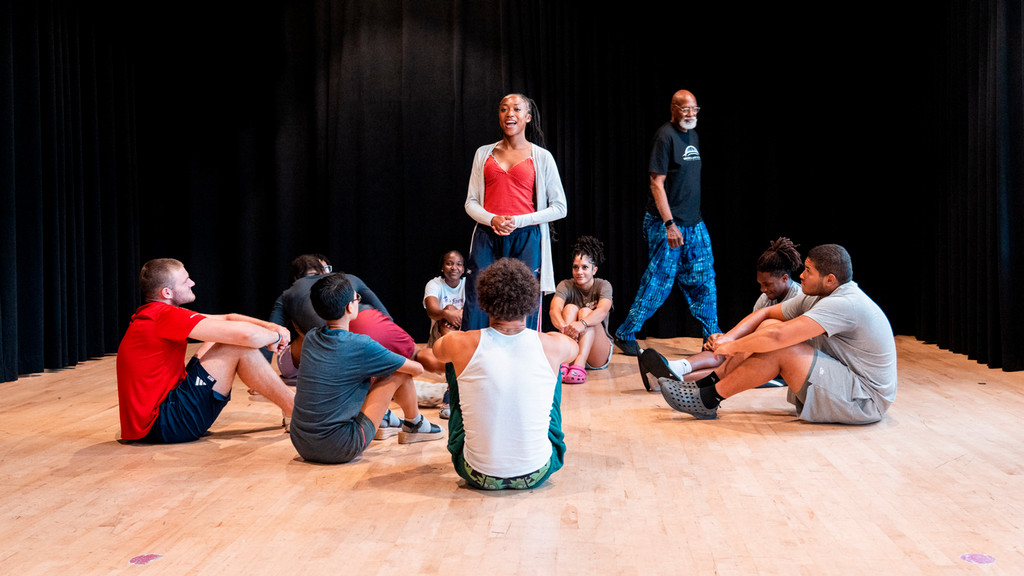 A theater class sits in a circle in a black box theater room, with one student standing in the center and a professor walking behind.