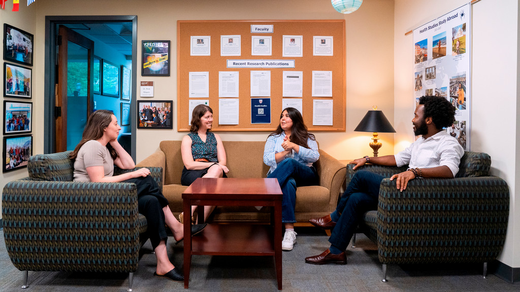 Four faculty members sit and talk together in a department lounge. 