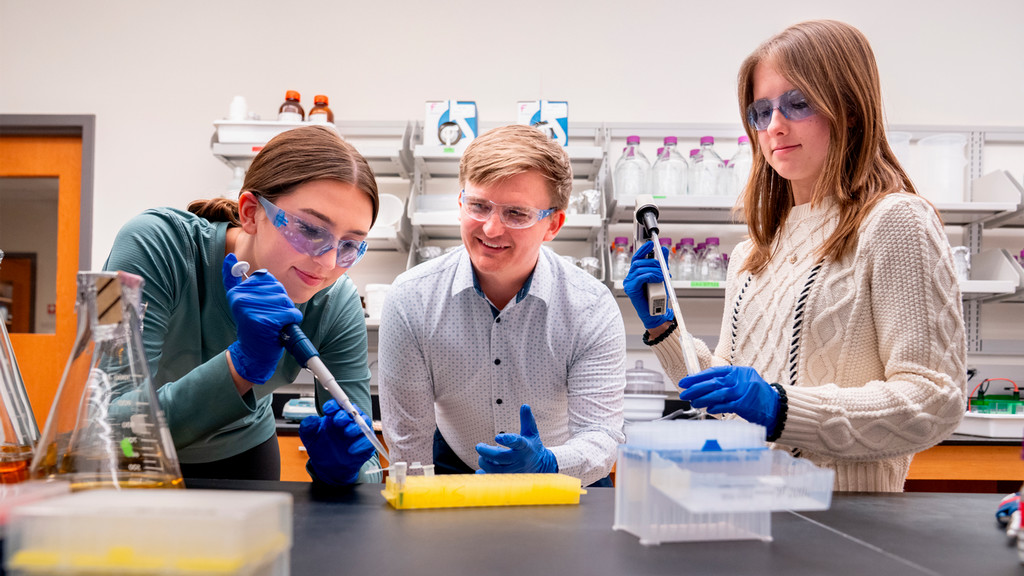 A professor works in a science lab with two students using pipettes.
