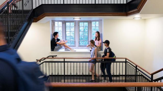 Interior of a residence hall. It is a large room with a high ceiling, windows on the far walls, and stairs going up and down to the various floors of the building.