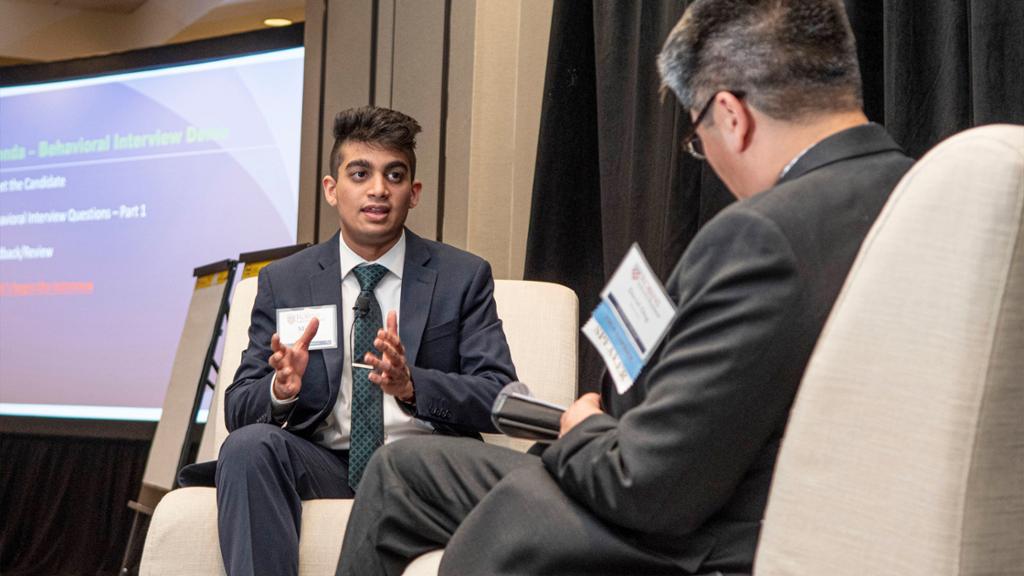 A student in a suit sits on stage talking with a keynote speaker.