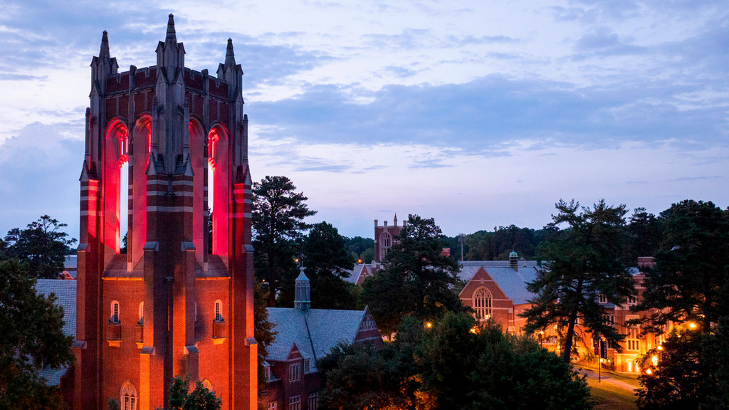 Brick gothic tower backlit in red against an evening sky, along with other lit campus buildings in the background