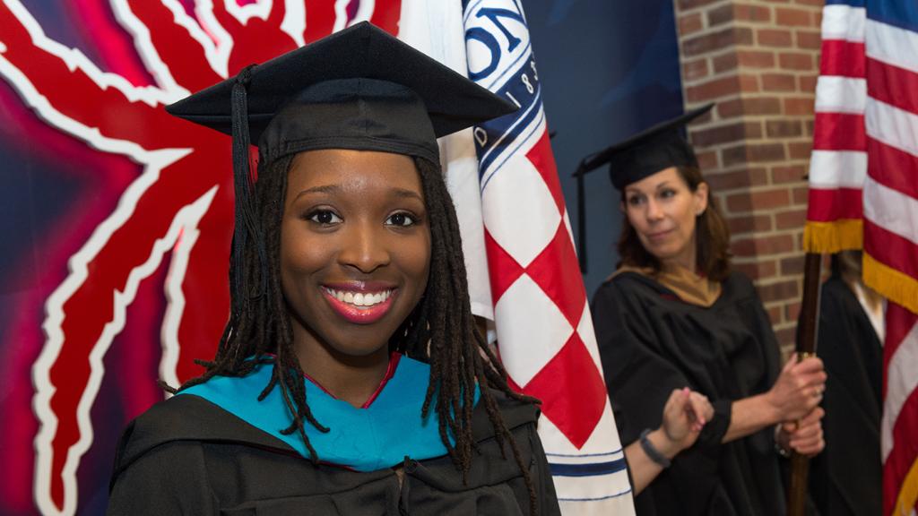 Student in cap and gown in front of flag and UR Spider