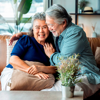 Happy senior Asian couple sitting together on a beige couch in a bright, modern living room, smiling and embracing while surrounded by potted plants and natural light from windows.