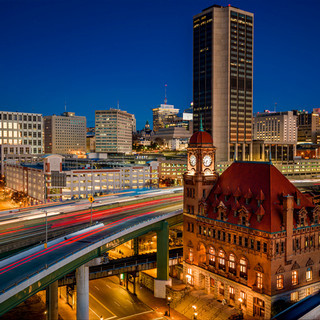 Historic Richmond, Virginia skyline at twilight featuring illuminated downtown skyscrapers, a grand red-roofed historic building with clock tower, elevated highway with light trails from traffic, and modern office buildings against a deep blue evening sky.