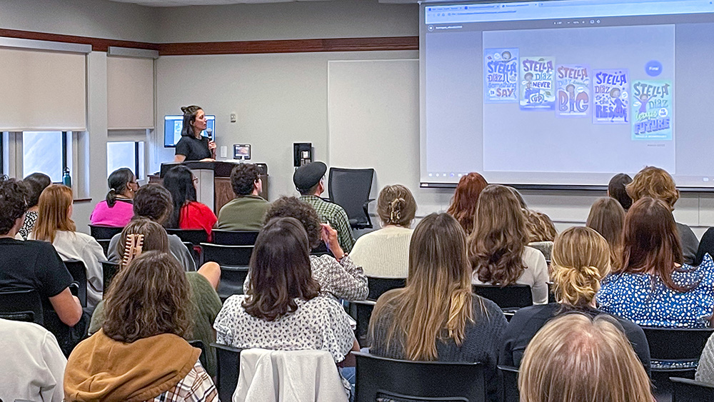 Author Angela Dominquez speaking to a group of listeners seating and looking at a screen showing book front covers