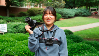 A student displays a camera while also wearing a GoPro camera while outside on campus.