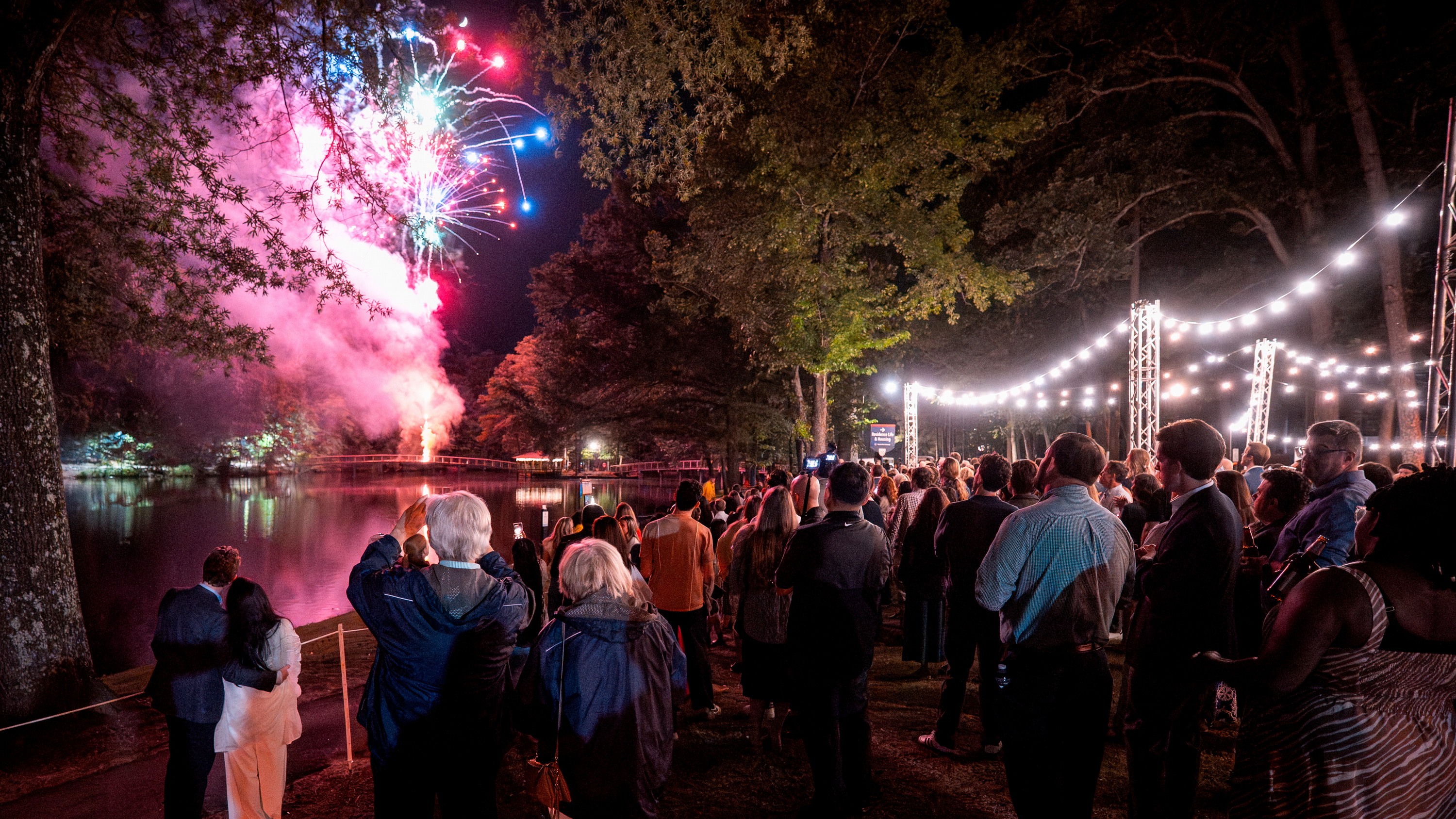 A photograph of  crowd of people watchign fireworks together over Westhampton Lake at the University of Richmond, shot from behind.
