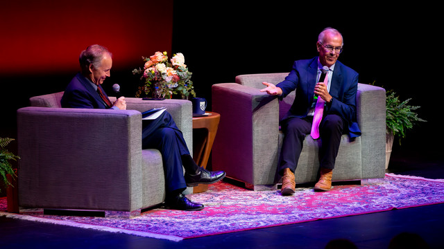 Writer David Brooks sitting on a chair on stage. He is talking with University chaplain Craig Kocher, and is holding a microphone and gesturing with his other hand.