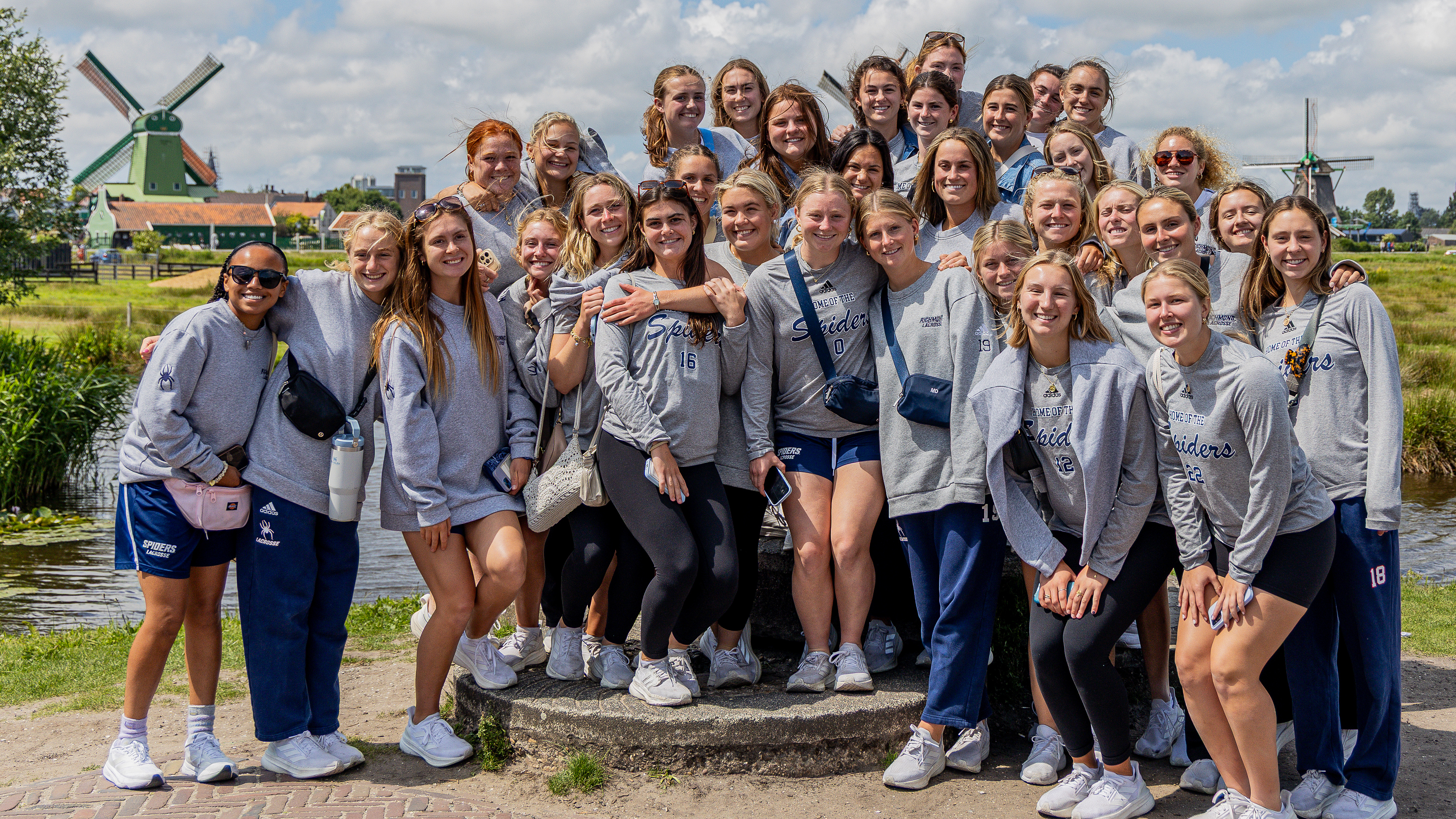 A photo of the women's lacrosse team posing for the camera. They are  in the Netherlands, and are standing outside, flanked by two windmills.
