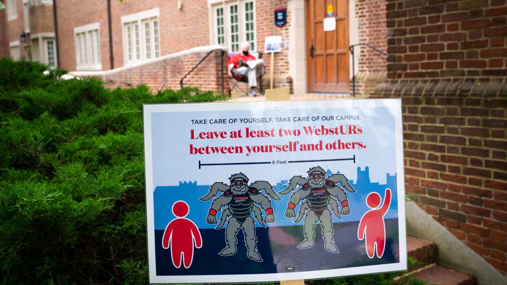 In the background, a man sits outside of a residence hall. In the foreground is a sign instructing people to maintain six feet of separation.