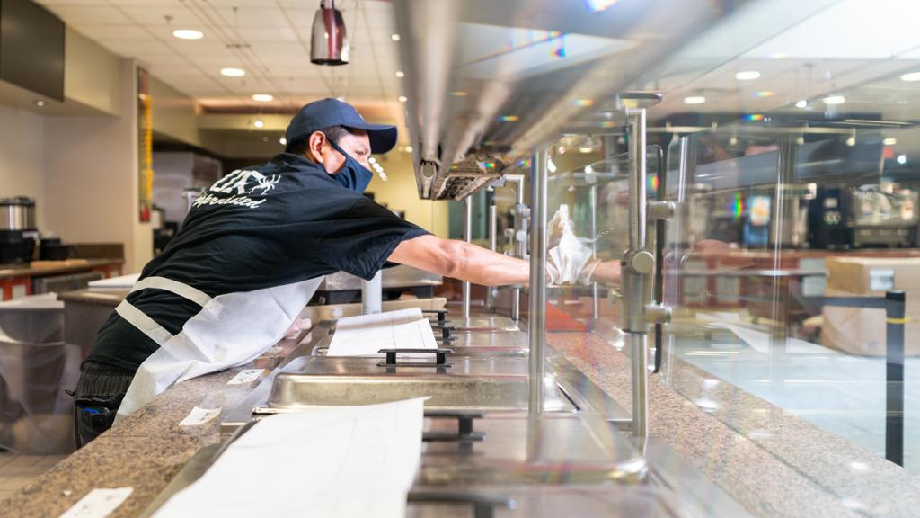 A dining hall employee wipes down a glass barrier inside the dining hall serving area.